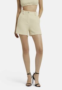 Beige geweven shorts met een hoge taille, voorzakken en knoopdetails, gecombineerd met zwarte enkelbandhaksandalen.