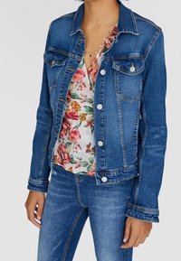 Chaqueta de mezclilla en azul medio con un corte clásico, botones plateados y bolsillos frontales. Lleva puesta sobre una blusa de estampado floral y combinada con jeans.