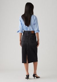 Black denim skirt with a back slit, featuring two back pockets and a leather patch at the waistband. Paired with a light blue shirt.