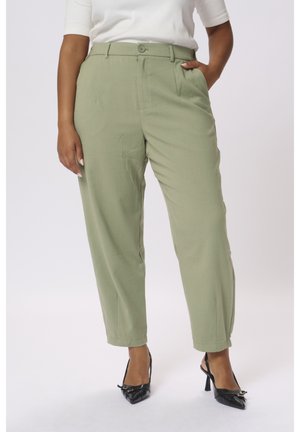Femme portant un pantalon taille haute vert clair, fuselé avec poches latérales, associé à un haut blanc et des escarpins slingback noirs à bout pointu.