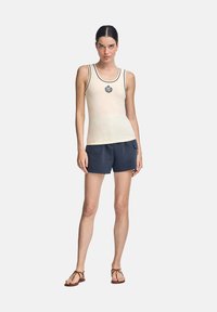 Top in beige con bordi neri e un emblema centrale, abbinato a pantaloni corti blu navy. Il top ha un design aderente e una texture liscia.