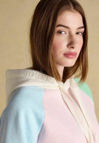 Jeune femme aux cheveux bruns lisses portant un sweat à capuche pastel avec des panneaux bleus, roses et verts, et le mot « JOULES » brodé sur la capuche.