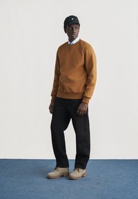 Άνδρας που φοράει καφέ φούτερ, μαύρο παντελόνι, μπεζ μπότες και μαύρο καπέλο, στέκεται σε μπλε χαλί μπροστά από λευκό τοίχο.