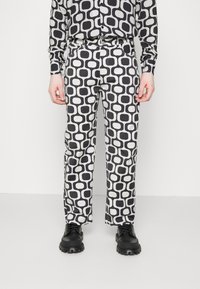 Person klädd i en matchande svart och vit geometrisk mönstrad skjorta och byxor med svarta kraftiga skor, stående mot en neutral bakgrund.