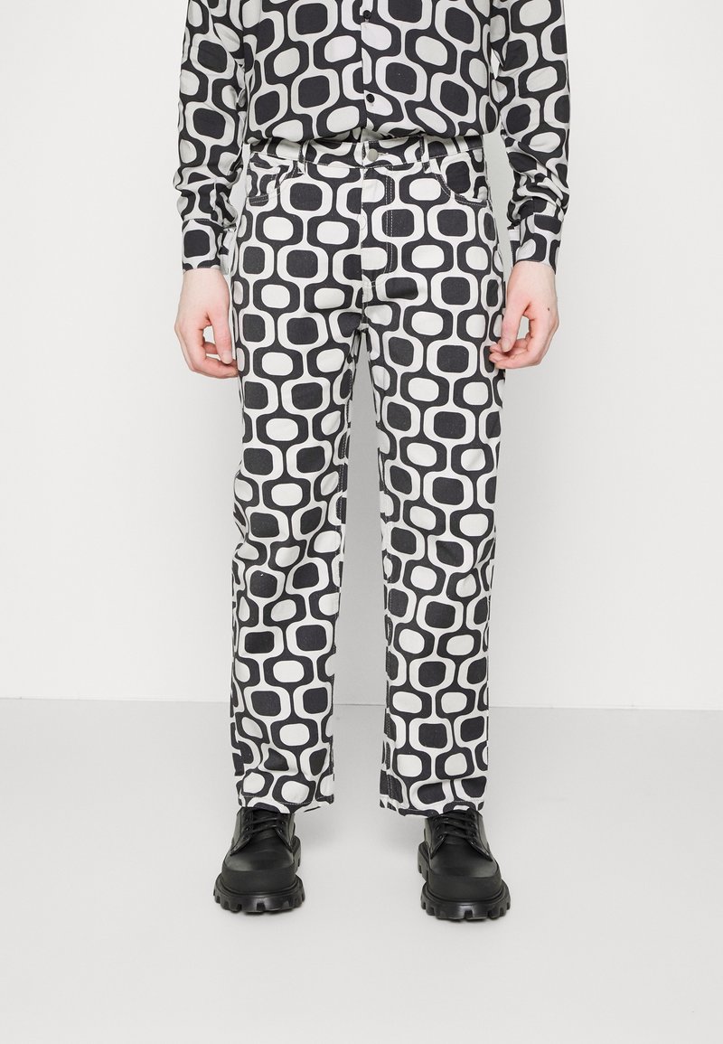 Person klädd i en matchande svart och vit geometrisk mönstrad skjorta och byxor med svarta kraftiga skor, stående mot en neutral bakgrund.