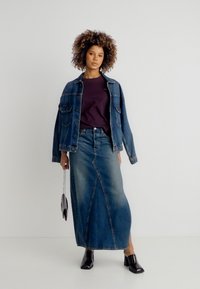 Γυναίκα που φοράει τζιν μπουφάν, σκούρο μωβ πουκάμισο, μακριά τζιν φούστα, μαύρα παπούτσια με τακούνι και κρατά μια μικρή τσάντα στο πλάι της.