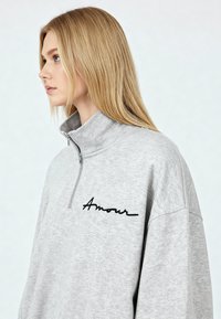 Sweat-shirt gris à fermeture éclair, doté d'un col montant et du mot « Amour » brodé en noir. Texture douce avec une coupe décontractée.