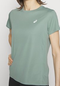 Světle zelené sportovní tričko z látky odvádějící vlhkost, s kulatým výstřihem, krátkými rukávy a malým bílým logem na hrudi.