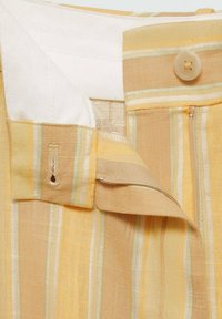 Geel en beige gestreepte broek met een tailleband, voorzien van een knoop, rits en zichtbare witte binnenvoering.
