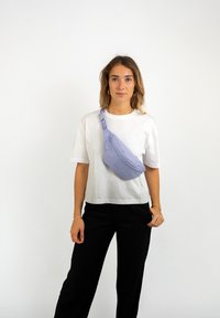 Sac bandoulière violet clair en forme courbée, doté d'une poche zippée à l'avant, d'une bandoulière réglable et d'une texture lisse. Porté sur un fond blanc.