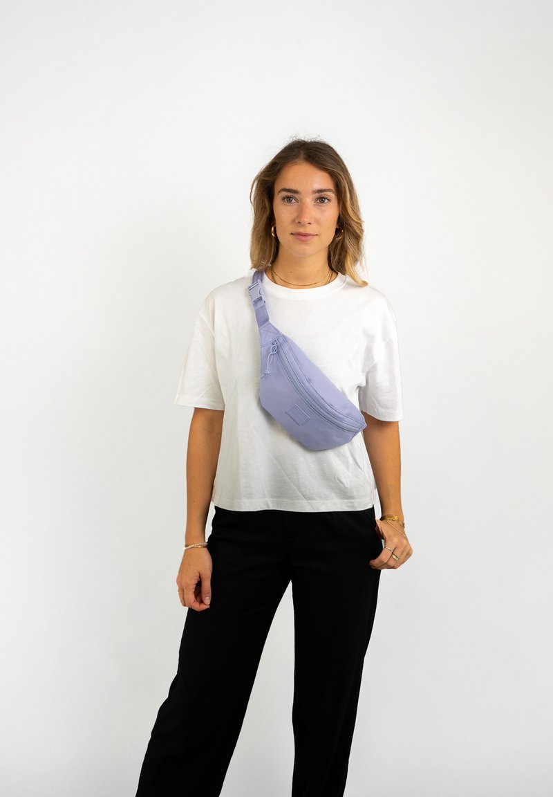 Sac bandoulière violet clair en forme courbée, doté d'une poche zippée à l'avant, d'une bandoulière réglable et d'une texture lisse. Porté sur un fond blanc.