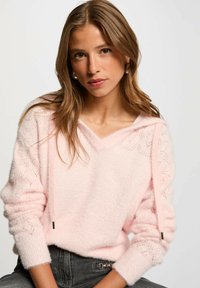 Maglione rosa chiaro con una texture morbida e pelosa, caratterizzato da una scollatura a V e motivi decorativi a maglia sulle maniche. Coulisse con punte in metallo.