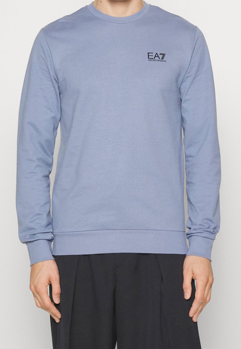 Homme portant un sweat-shirt EA7 Emporio Armani bleu clair à manches longues avec un pantalon noir, debout devant un fond uni.