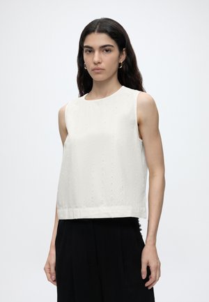 Γυναίκα με μακριά σκουρόχρωμα μαλλιά φοράει λευκό αμάνικο τοπ με τρυπητό μοτίβο και μαύρο παντελόνι, στέκεται μπροστά σε απλό ανοιχτό φόντο.