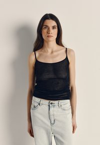 Jauna sieviete ar gariem tumšiem matiem, tērpusies melnā bezroku topā un gaiši zilās džinsās, stāv uz vienkārša bēša fona.