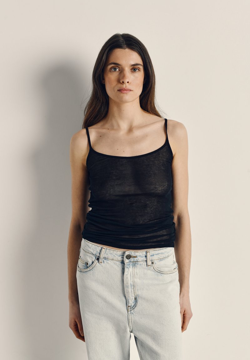 Jauna sieviete ar gariem tumšiem matiem, tērpusies melnā bezroku topā un gaiši zilās džinsās, stāv uz vienkārša bēša fona.