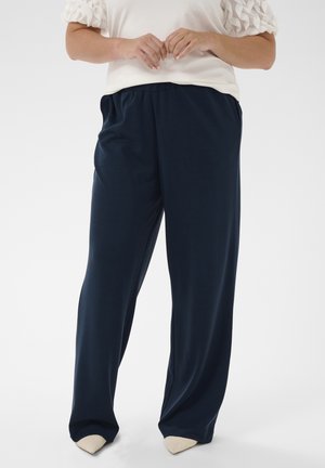 Donna con indosso un top bianco con maniche a sbuffo e pantaloni larghi blu scuro, in piedi con le mani intrecciate davanti.