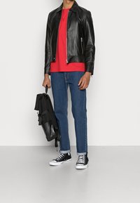 Uomo che indossa una giacca di pelle nera, una camicia rossa, jeans blu e sneakers nere, mentre tiene uno zaino nero a lato contro uno sfondo semplice.