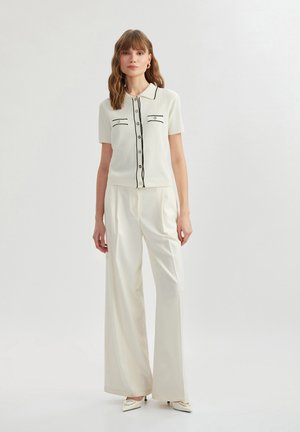 Polo shirt bianco a coste con rifiniture nere, maniche corte e due tasche frontali, abbinato a pantaloni bianchi a gamba larga e scarpe con punta affusolata.