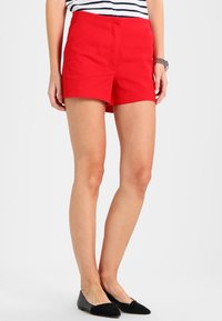 Shorts rouges à texture lisse, devant plat et poches. Associés à un haut rayé et des ballerines noires.