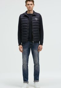 Gilet imbottito nero con colletto alto, chiusura con zip e logo sul petto, abbinato a una maglietta scura a maniche lunghe, jeans blu e sneakers bianche.