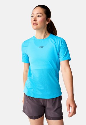 Vrouw met opgestoken haar, gekleed in een lichtblauw Brooks hardloopshirt en donkergrijze gelaagde korte broek, die licht omhoog en naar links kijkt.