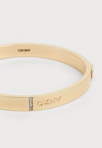 Kuldne metallist käevõru, millel on lame disain, graveeritud "DKNY" ja väike rida selgeid kristalle rõhutamiseks.