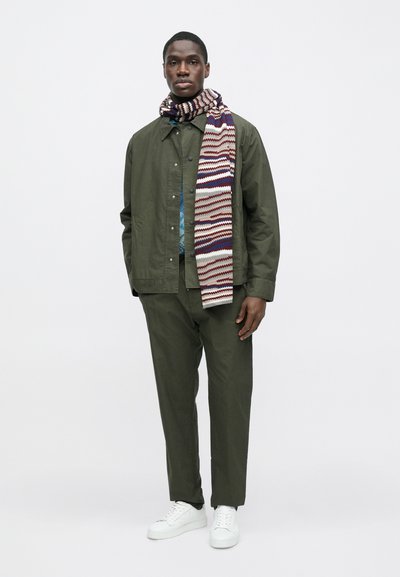 Χακί πράσινο μπουφάν και παντελόνι σε συνδυασμό με πολύχρωμο πλεκτό κασκόλ. Λευκά αθλητικά παπούτσια ολοκληρώνουν την εμφάνιση, με άνετη εφαρμογή και casual σχεδιασμό.