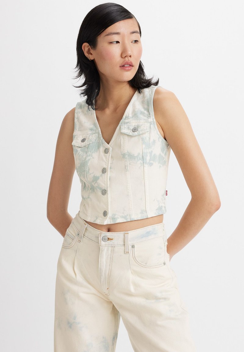 Cropped denim vest in cream with light blue tie-dye patterns. Features a V-neck, button closure, and two chest pockets. Smooth texture.
