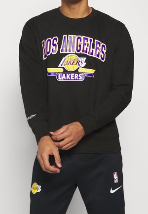 Person trägt schwarzen Los Angeles Lakers Sweatshirt und passende schwarze Hose mit NBA- und Nike-Logos, Hände nahe der Taille.