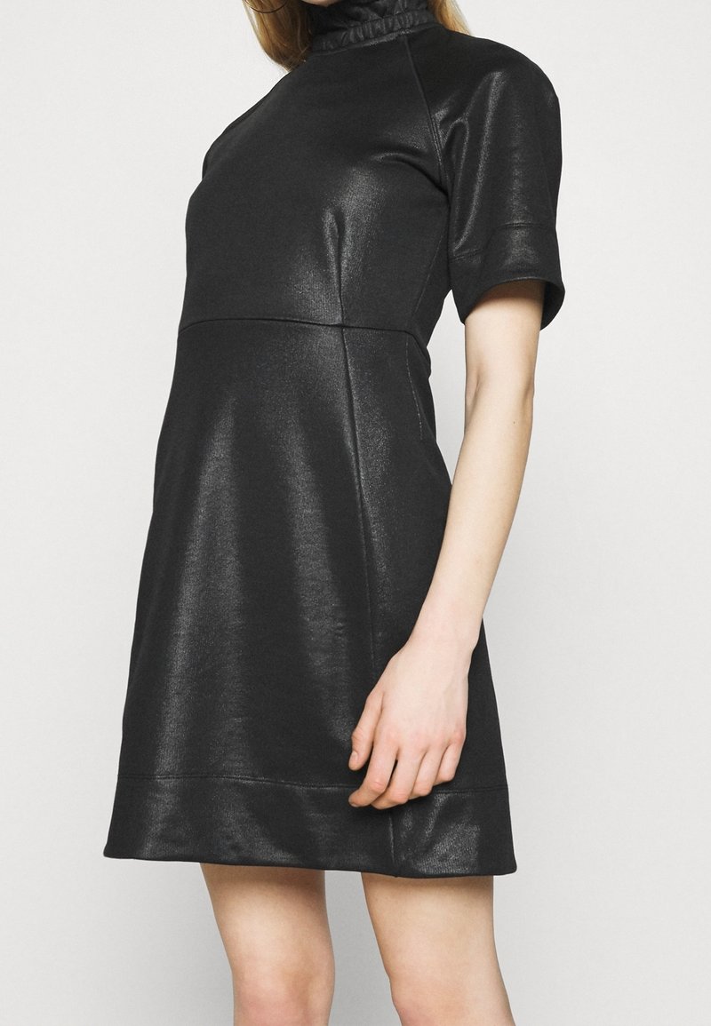 Femme portant une robe noire brillante à manches courtes avec un col montant à volants, debout devant un fond clair uni.
