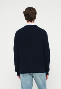 Granatowy sweter z dzianiny z okrągłym dekoltem, ściętym hemem i długimi rękawami, noszony na jasnej koszuli z kołnierzykiem, zestawiony z jasnoniebieskimi dżinsami.