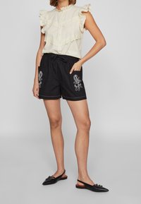Blouse crème avec des manches froncées et des détails en dentelle, accompagnée de shorts noirs brodés avec un cordon de serrage et des poches latérales. Chaussures noires.