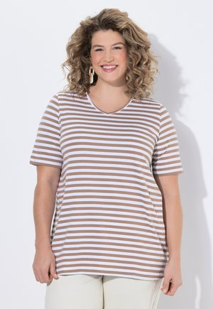 Lächelnde Frau mit lockigem Haar, die ein beige-weiß gestreiftes kurzärmeliges Hemd und hellfarbene Hosen vor einfachem Hintergrund trägt.