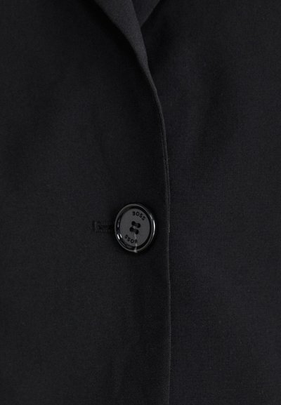 Blazer noir avec un tissu texturé, arborant un bouton noir proéminent portant l'inscription "BOSS" et un design de revers épuré et simple.