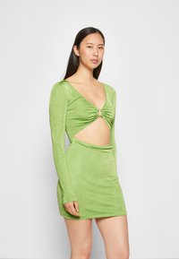 Vestido verde de mangas largas com uma silhueta ajustada, apresentando um design recortado na cintura, drapeado e um detalhe metálico circular na gola.