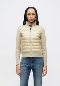 Μπεζ ζακέτα με φερμουάρ, με επένδυση στο μπροστινό μέρος, ριμπ μανσέτες και γιακά. Διαθέτει οριζόντιες ραφές και λογότυπο στο στήθος. Φορεμένη με μπλε τζιν.