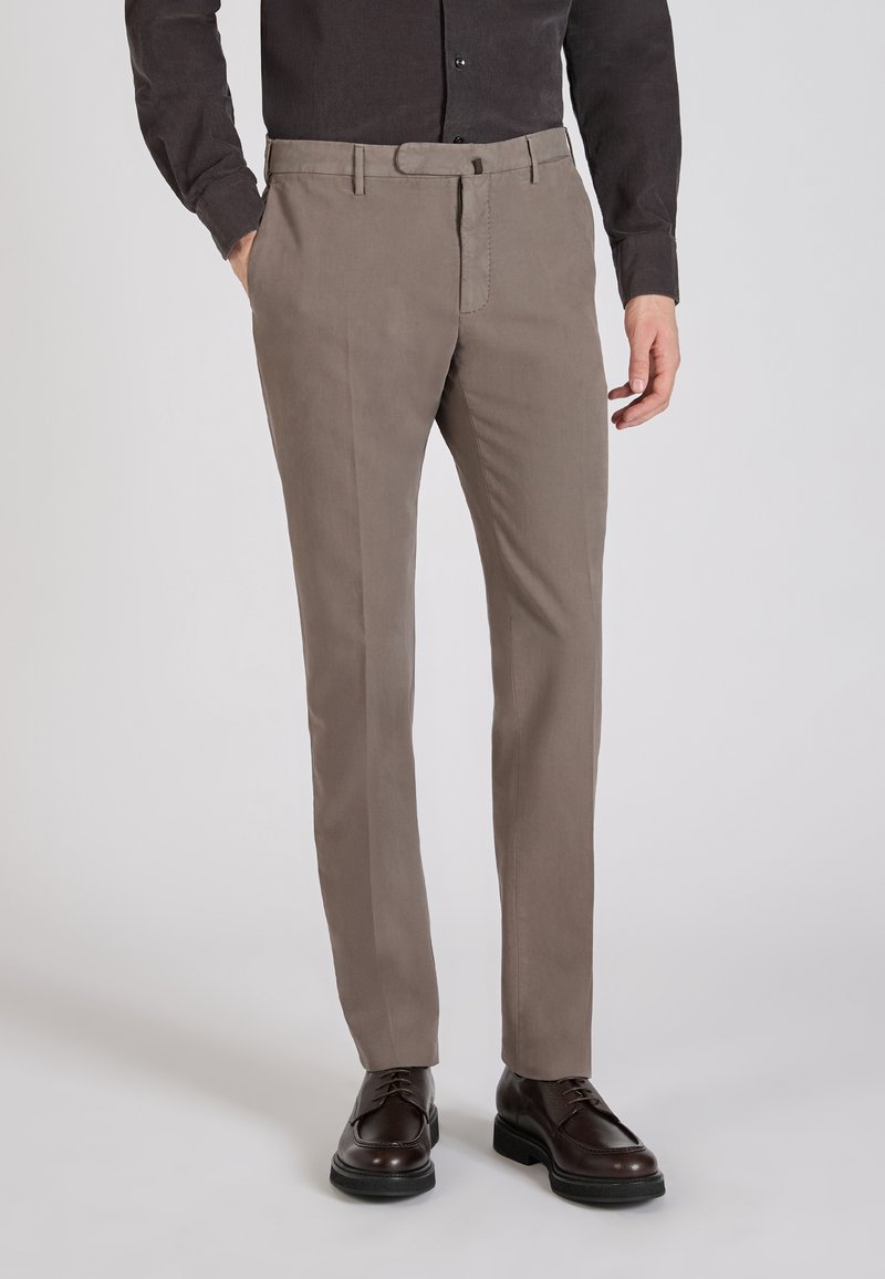 Brązowe spodnie o dopasowanym kroju, wykonane z lekkiego materiału, z płaskim przodem, paskami na pasek i zwężającymi się nogawkami, zestawione z ciemnymi butami.