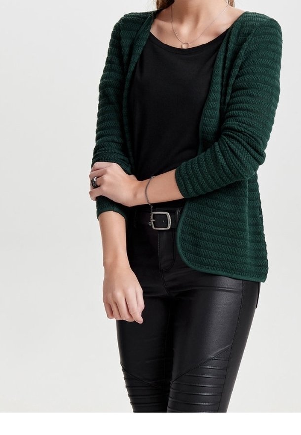 Femme portant un cardigan texturé vert foncé par-dessus un haut noir et un pantalon en cuir noir, les bras croisés à la taille.