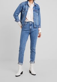 Persona con jeans azul de tiro alto y puños, chaqueta de mezclilla a juego, camisa blanca con patrón y botas blancas hasta el tobillo, de pie sobre un suelo gris.