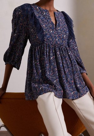 Haut floral en tissu bleu marine avec manches ballon, détails en dentelle sur les épaules et taille froncée. Associé à un pantalon de couleur claire.