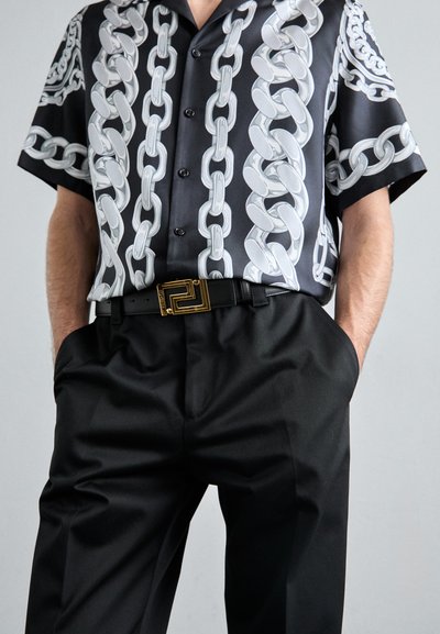 Camisa negra con un patrón de cadena brillante, mangas cortas y botones, combinada con pantalones negros ajustados y un cinturón negro con detalles en dorado.
