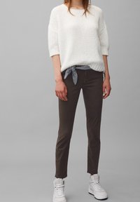 Maglione bianco lavorato a maglia con maniche corte, abbinato a pantaloni marroni aderenti con una cintura a cravatta decorata. Il modello indossa sneakers bianche.