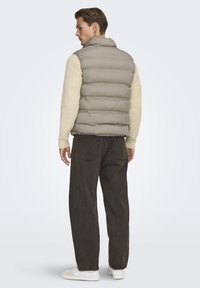 Gilet imbottito beige chiaro con colletto alto, abbinato a un maglione a coste crema e jeans marroni scuri, con una vestibilità rilassata e sneakers bianche.