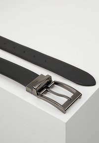 Cinturón de cuero negro texturizado con una hebilla rectangular de metal plateado, que presenta un diseño clásico y agujeros perforados para el ajuste.