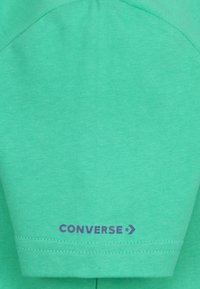 Vaaleanvihreä, lyhythihainen t-paita, joka on valmistettu puuvillasta. Siinä on violetti "CONVERSE" -logo ja tähtiakseli helman lähellä. Sileä rakenne.
