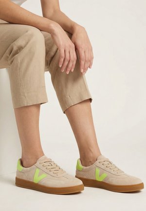 Person iført beige korte bukser og beige sneakers med neon grønne accenter, siddende med hænder hvilende på knæene.