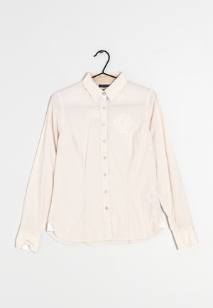 Chemise beige à manches longues avec boutons, fines rayures verticales et col, affichée sur un cintre noir sur fond blanc.