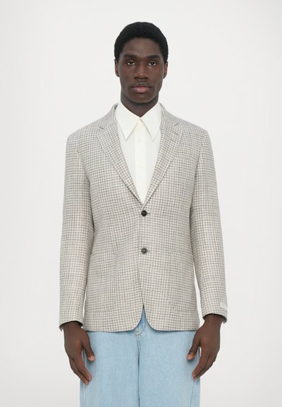 Szürke hounds-tooth mintás blazer, két gombbal, hajtókás gallérral és egyetlen mellzsebbel, fehér galléros inggel és világoskék farmernadrággal párosítva.