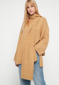 Kaabla kudumiga kõrge kaelusega kampsun helepruunis toonis, millel on pikk sallilaadne detail, pikendatud varrukad ja lõdva istuvusega. Kandmiseks siniste teksadega.